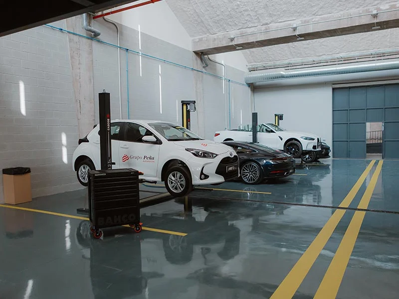 Tres coches aparcados en el interior de un limpio y moderno taller de reparación de automóviles MOTIO, con suelos relucientes y un carro de herramientas cerca, listo para la temporada del curso 2025-2026.