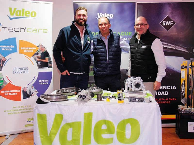 Tres hombres de pie detrás de una mesa en la que se exponen piezas de automóviles y una pancarta de Valeo en un evento automovilístico, mostrando "un paso de gigante" en la innovación automovilística.