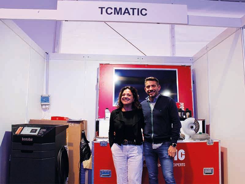 Dos personas de pie y sonrientes delante de un expositor rojo con la etiqueta "TCMATIC", celebrando un paso de gigante en su viaje juntos.
