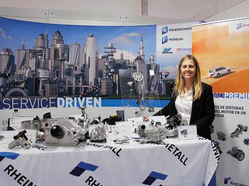 Una mujer junto a un expositor de piezas de motores de automóvil en un stand con un paisaje urbano de fondo, que pone de relieve un verdadero paso de gigante de la ingeniería moderna.