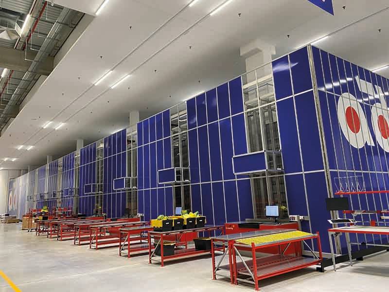 Filas de puestos de trabajo industriales azules con mesas y ordenadores rojos se alinean en una fábrica espaciosa y bien iluminada: un verdadero paso de gigante en la eficiencia de la fabricación moderna.