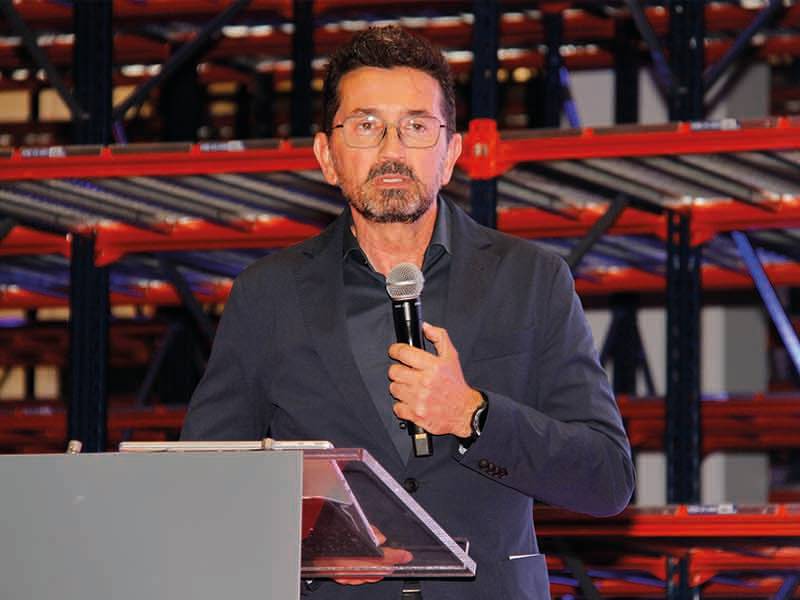 Un hombre con gafas habla por un micrófono en un podio, dando un paso de gigante mientras unas estanterías metálicas rojas se alzan en el fondo.