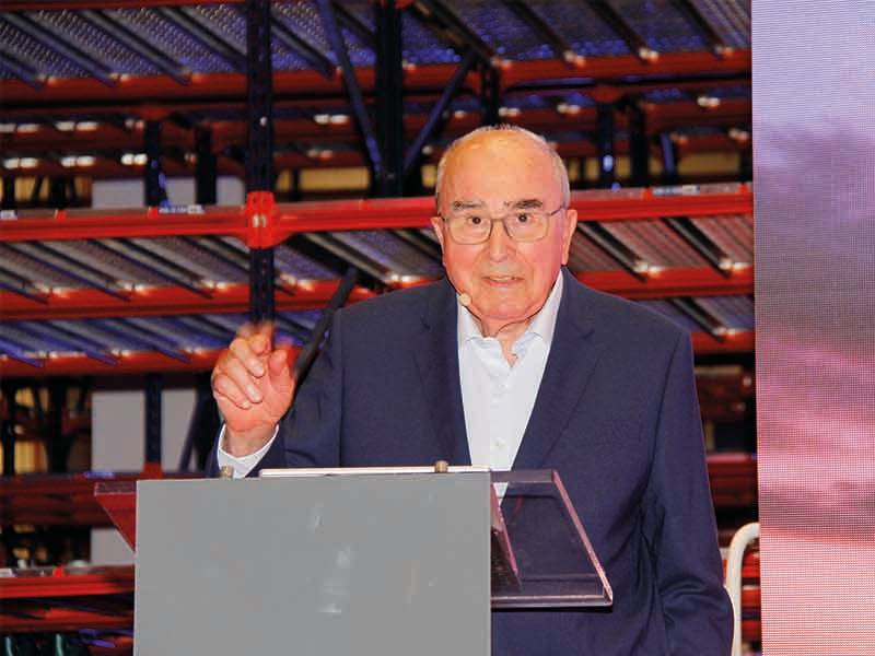 Anciano trajeado hablando en un podio con estanterías industriales al fondo, marcando un verdadero paso de gigante para la empresa.