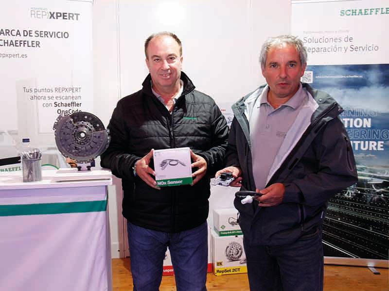 Dos hombres de pie en el interior de un stand promocional, sosteniendo piezas de automóviles y una caja de productos, mostrando un paso de gigante en soluciones innovadoras para automóviles.
