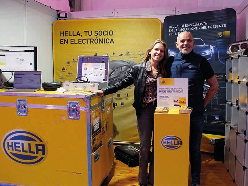Dos personas sonríen en un stand de electrónica de HELLA, con vitrinas amarillas y material promocional, para celebrar un paso de gigante en la innovación automovilística.