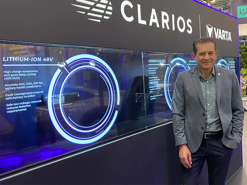 Un hombre de traje gris junto a un expositor de Clarios y Varta con información sobre baterías de iones de litio y soluciones de movilidad, en el que también aparecen productos de Optima.