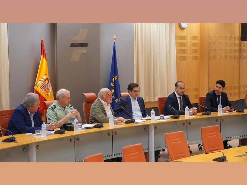 Seis hombres sentados en una mesa de conferencias con las banderas de España y de la UE de fondo, enfrascados en una discusión.