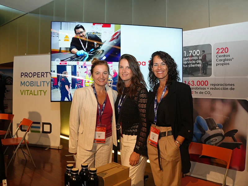 Tres mujeres en traje de negocios sonríen en un stand de un evento con pancartas promocionales y una gran pantalla de vídeo detrás de ellas.
