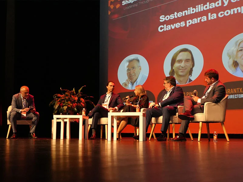 Cinco personas en traje de negocios se sientan en el escenario, hablando durante un panel de debate sobre sostenibilidad con un telón de fondo rojo.
