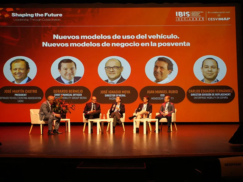Cinco hombres se sientan en el escenario con micrófonos en un panel de una conferencia, con los nombres de los oradores mostrados en una pantalla detrás de ellos.