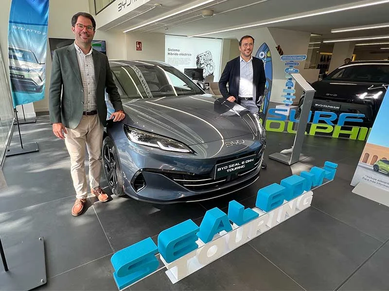 Dos hombres junto a un coche BYD Seal gris en una sala de exposición con expositores azules Touring y Surf Dolphin.