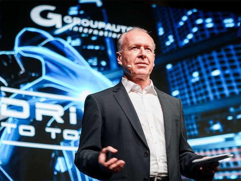 Un hombre trajeado habla en el escenario con gráficos futuristas y la inscripción "GROUPAUTO 2025 Supplier Global Meeting" a sus espaldas.