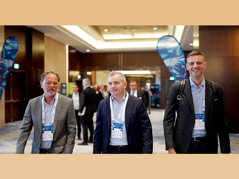 Tres hombres en traje de negocios caminan juntos en la Reunión Mundial de Proveedores 2025, portando insignias, en un moderno local interior organizado por GROUPAUTO.
