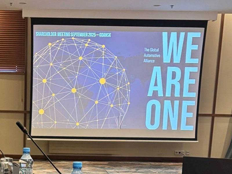 Pantalla de proyección con el lema "WE ARE ONE" y un gráfico de la red mundial en la reunión de accionistas de Global One Automotive en Gdansk, septiembre de 2025.