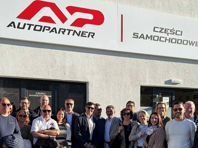 Un grupo de personas sonríe en el exterior de un edificio de AutoPartner durante una reunión de accionistas bajo un cartel con texto en polaco, en representación de Global One Automotive.
