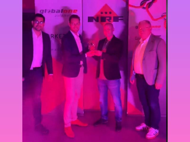 Cuatro hombres en traje de negocios permanecen juntos en una reunión de Global One Automotive, rodeados de pancartas e iluminación rosa.