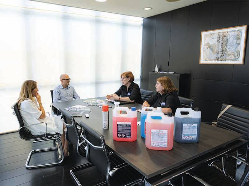 Cuatro personas sentadas en una mesa de conferencias con grandes recipientes de productos químicos de limpieza -cada uno de ellos etiquetado como aumentar capacidad- colocados en primer plano.