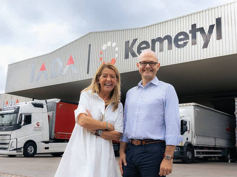 Dos personas sonríen delante de un edificio con un cartel de "Kemetyl" y varios camiones grandes aparcados cerca, lo que refleja los esfuerzos por aumentar la capacidad y la eficiencia.