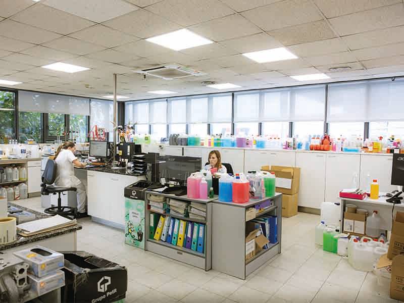 Dos personas trabajan en un luminoso laboratorio lleno de botellas de colores, equipos y papeleo, colaborando para aumentar la capacidad de producción hasta en un 60%.
