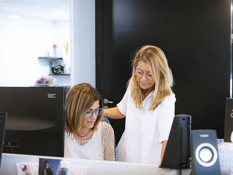 Dos mujeres en una oficina, una sentada ante un ordenador y la otra de pie a su lado, ambas mirando la pantalla mientras discuten cómo aumentar la capacidad en un 60%.