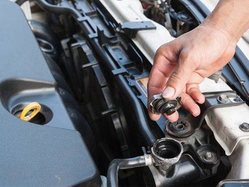 Una mano retira el tapón del radiador del motor de un coche, con el capó abierto para su mantenimiento, un paso esencial para preparar tu vehículo para el invierno con productos térmicos y soporte técnico especializado.