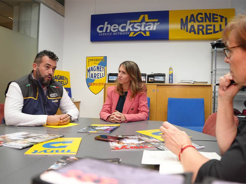 Tres personas conversan sentadas a una mesa en una oficina, con carteles de Magneti Marelli Parts & Services en la pared a sus espaldas, que ponen de manifiesto su cercanía al cliente.
