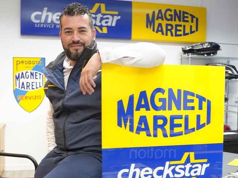 Hombre sentado y sonriente cerca de un gran cartel de Magneti Marelli en una oficina con carteles de la marca en las paredes, lo que refleja un ambiente acogedor centrado en el servicio al cliente y las piezas de automóviles de calidad.