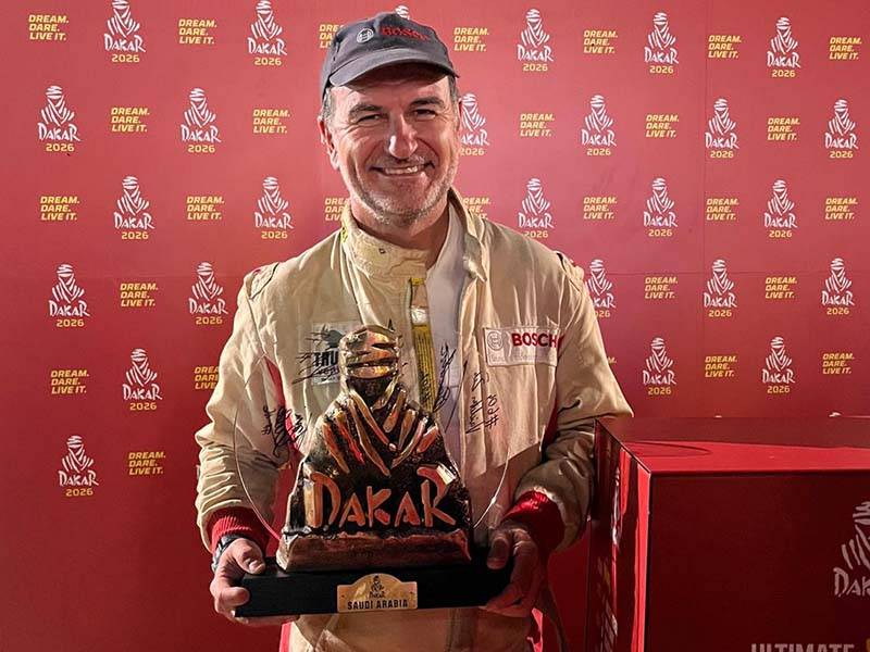 Hombre sonriente con equipo de carreras sosteniendo un trofeo del Dakar, de pie delante de un telón de fondo del Rally Dakar 2026.
