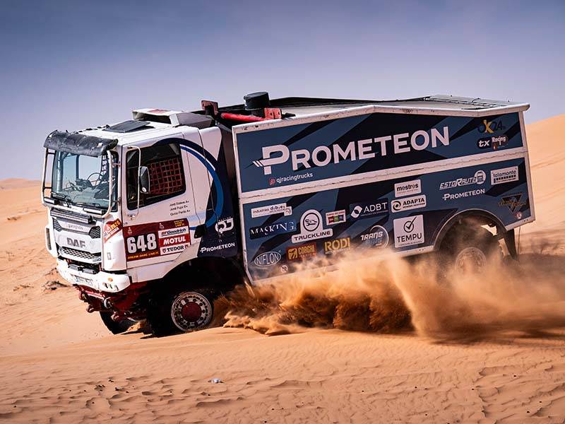 Un gran camión de rally El Dakar corre por la arena del desierto a lo largo de un desafiante sector, levantando polvo; está cubierto de logotipos de patrocinadores y lleva el número 648.