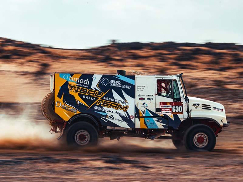 Un camión de rally con coloridos logotipos de patrocinadores atraviesa a toda velocidad un sector desértico del recorrido de El Dakar, levantando polvo tras de sí.