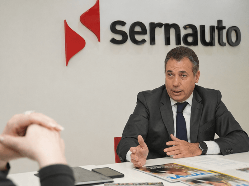 Hombre trajeado hablando en una mesa con el logotipo de Sernauto en la pared detrás de él.