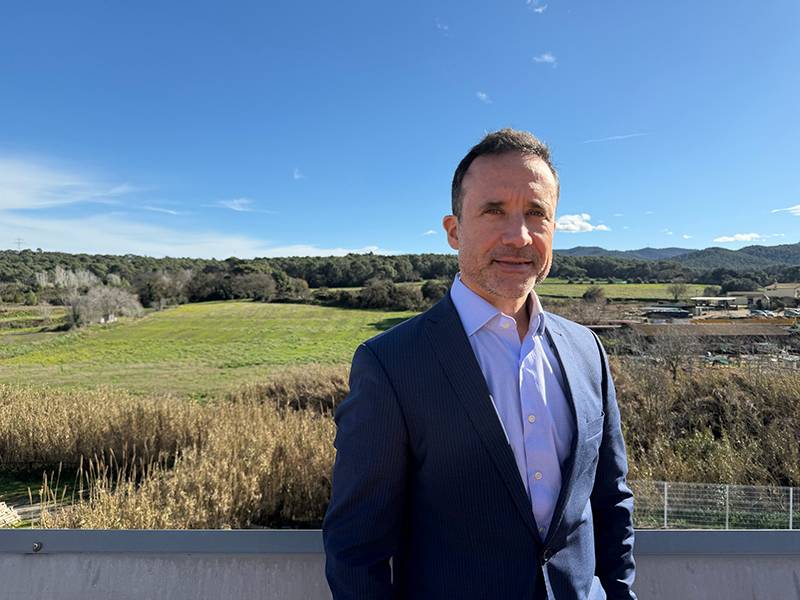 Hombre con traje azul de pie al aire libre con campos verdes, árboles y un cielo azul de fondo.