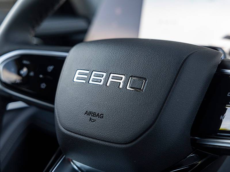 Detalle del volante de un coche de lujo Ebro S700 con el logotipo "EBRO" y una etiqueta de airbag en el centro, que destaca su diseño premium PHEV.
