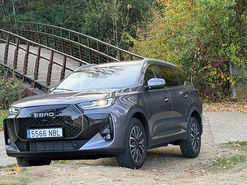 Un SUV Ebro S700 PHEV gris oscuro aparcado cerca de un puente de madera, rodeado de árboles y follaje otoñal.