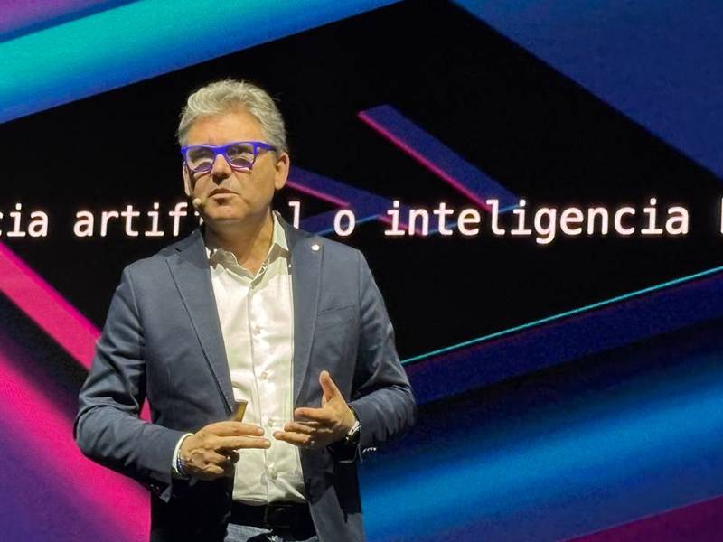Un hombre trajeado habla en el escenario con el texto "inteligencia artificial" y gráficos de neón de fondo.