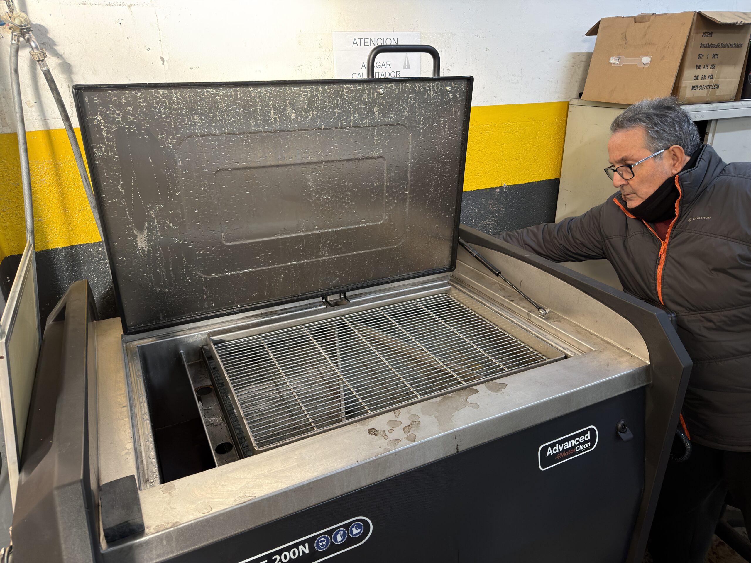 Un hombre con gafas mira dentro de una máquina industrial abierta con una rejilla metálica en un taller.