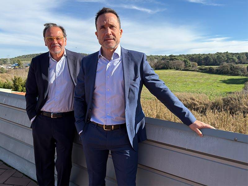 Dos hombres trajeados permanecen al aire libre junto a una barandilla con un campo de hierba y árboles al fondo bajo un cielo azul.