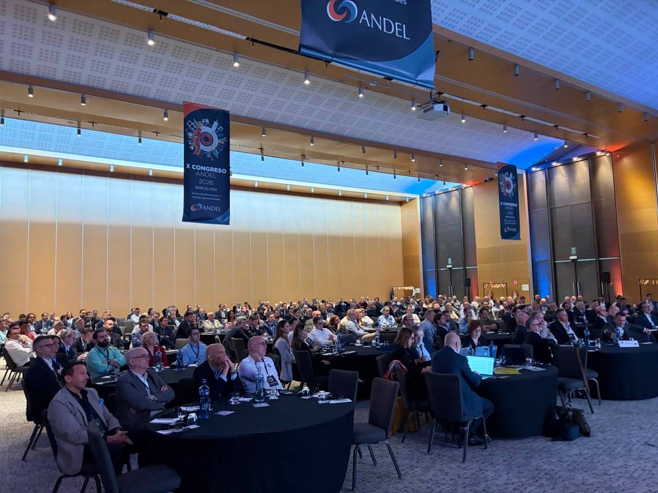 Gran grupo de personas sentadas en mesas redondas en una sala de conferencias, escuchando una presentación.