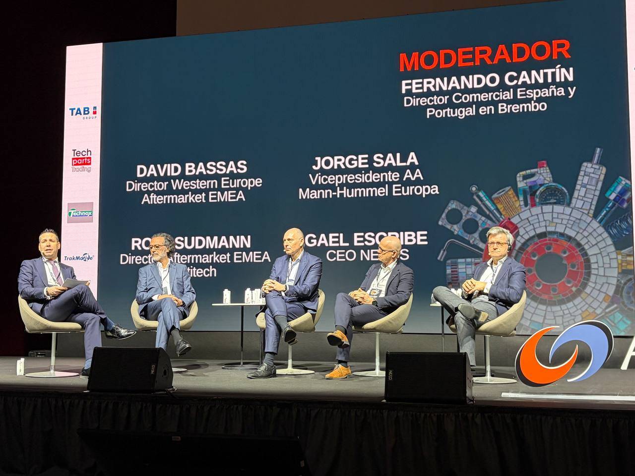 Cinco hombres trajeados se sientan en el panel del escenario con nombres y títulos que aparecen en una gran pantalla detrás de ellos.