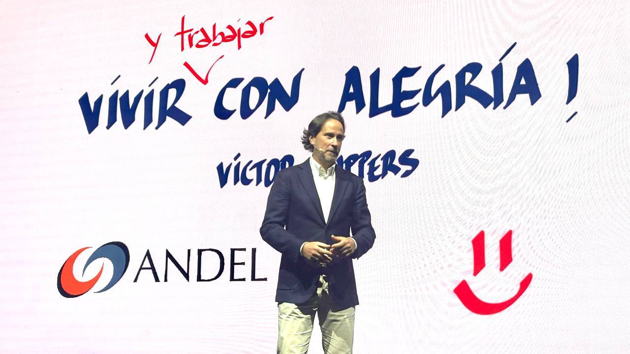 Un hombre sube al escenario frente a una pantalla en la que se lee "Vivir con alegría y trabajar" con el logotipo de ANDEL y una cara sonriente.