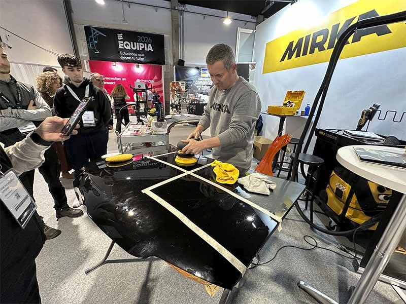 Un hombre hace una demostración de pulido de superficies de coches en el stand de una feria, atrayendo la asistencia de curiosos asistentes mientras muestra técnicas para lograr un acabado impecable.