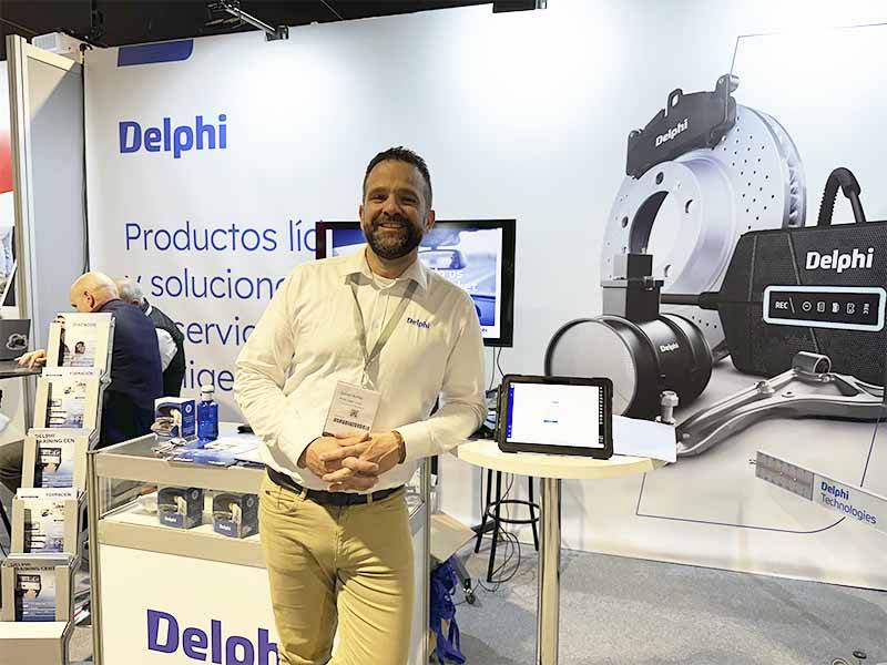Un hombre sonriente en un stand de Delphi en el que se exponen productos de automoción y se comparte la oferta de asistencia como parte de Balance de Equipia 2026 en una exposición.