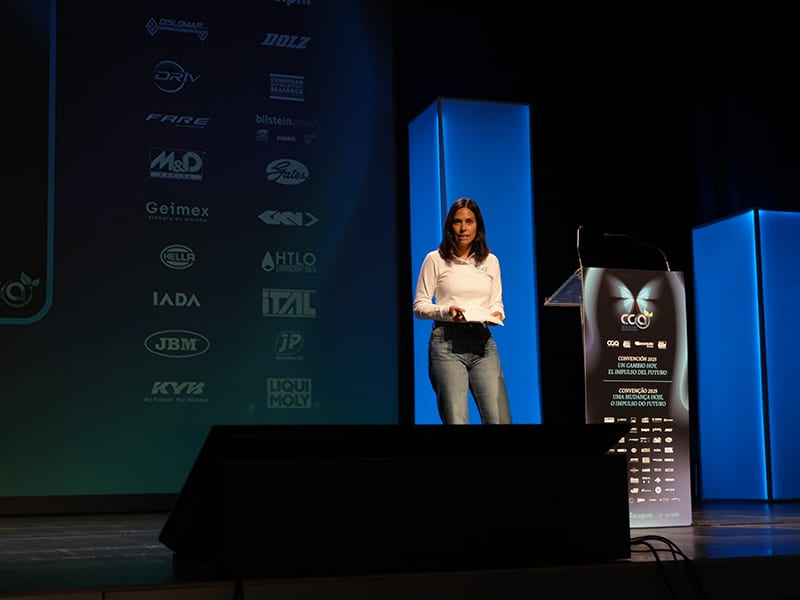 Una mujer con top blanco y vaqueros habla en el escenario de una conferencia, de pie cerca de un podio con logotipos de patrocinadores.