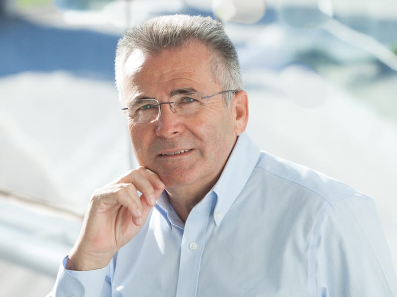 Un hombre mayor, con gafas y pelo canoso, vestido con una camisa azul claro, sonríe y apoya la barbilla en la mano.