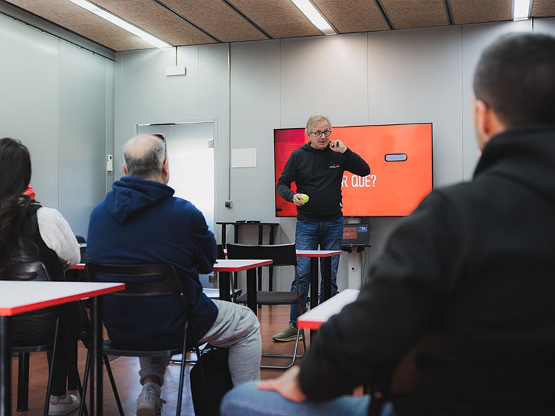 Un hombre presenta delante de un aula en Castellolí, dirigiendo un curso de conducción con una pantalla que muestra texto a las personas sentadas, centrándose en la prevención y las prácticas de conducción segura.