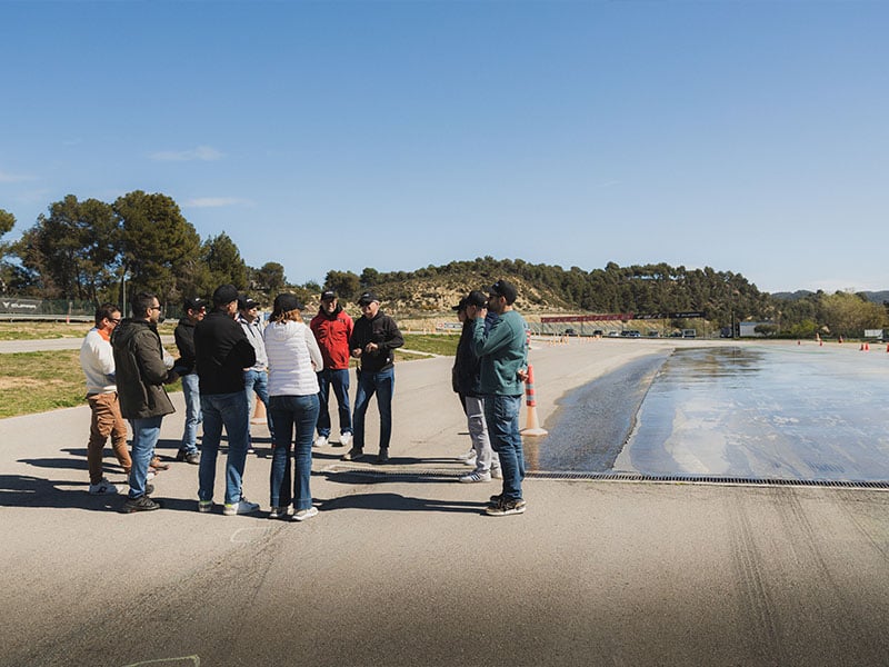 Un grupo de personas conversa cerca de una pista mojada al aire libre en un día soleado en Castellolí, con árboles y colinas de fondo durante un curso de conducción centrado en la prevención en carretera.