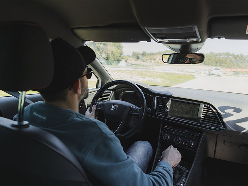 Hombre con gafas de sol y gorra conduciendo un coche tras realizar un curso de conducción en Castellolí, visto desde el asiento trasero en un día soleado.