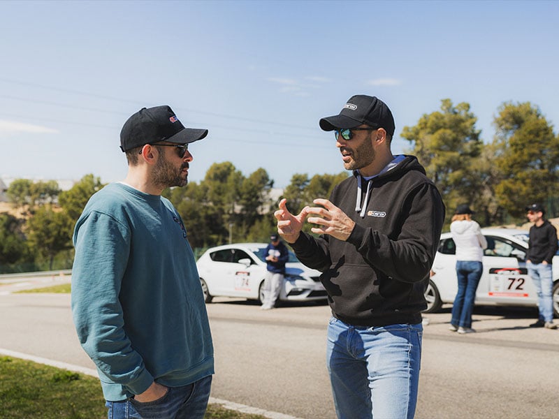 Dos hombres con ropa informal y sombreros hablan al aire libre cerca de coches blancos aparcados en un día soleado en Castellolí, posiblemente discutiendo curso de conducción o prevención carretera.