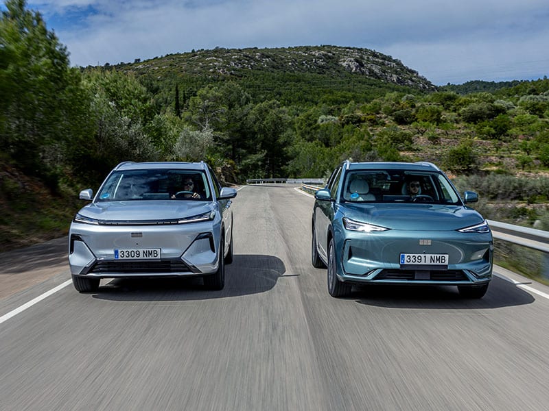 Dos SUV eléctricos, el Geely Starray EM-i y el E5 eléctrico, circulan uno al lado del otro por una carretera panorámica con colinas y exuberante vegetación de fondo.