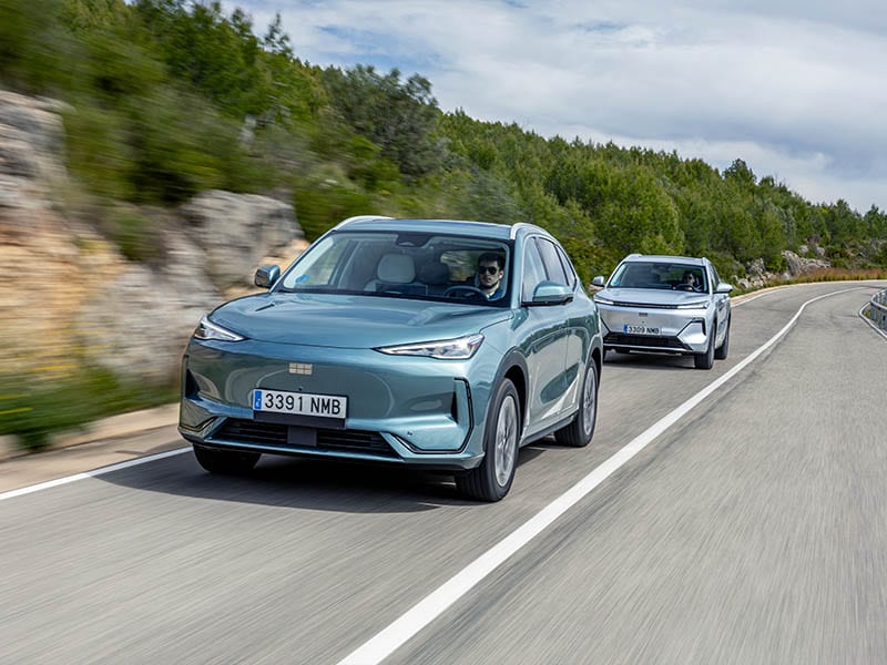 Dos todoterrenos eléctricos, el Geely E5 eléctrico y el Starray EM-i, circulan por una carretera sinuosa rodeada de árboles y colinas rocosas bajo un cielo nublado.